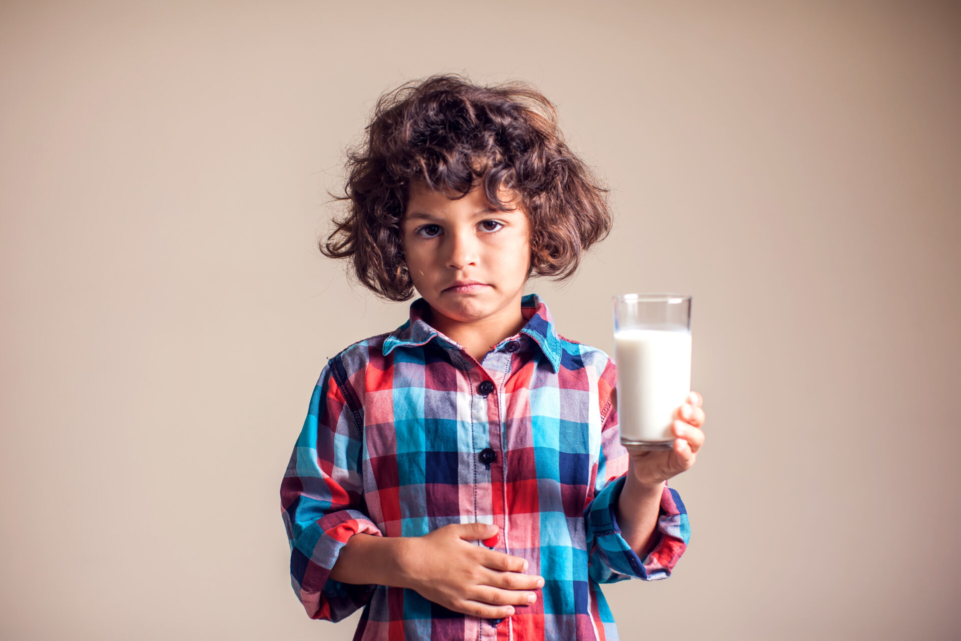 Child in a plaid shirt standing in front of a yellow backdrop holding a glass of milk in his right hand and clutching is stomach in pain with his left hand.