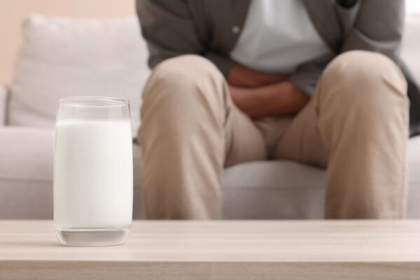 Featured image - a glass of milk is on the coffee table in the foreground while a man is sitting on the couch clutching his stomach in pain in the background.